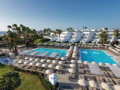 Hotelcomplex met twee grote zwembaden, parasols en palmbomen onder een heldere lucht.