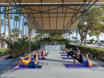 Groep mensen doet buiten yoga onder een groot afdak naast palmbomen.
