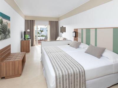 Camera d'hotel luminosa con due letti matrimoniali, TV e accesso al balcone.