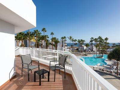 Balkon mit zwei Stühlen und Tisch, Blick auf Swimmingpool, Palmen und blauen Himmel.
