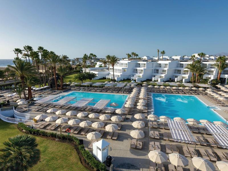 Complesso alberghiero con grandi piscine, lettini e palme sotto un cielo sereno vicino al mare.