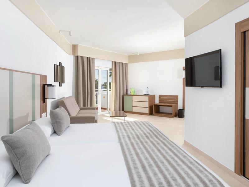 Camera d'hotel luminosa con due letti, cuscini grigi, TV a parete e accesso al balcone.