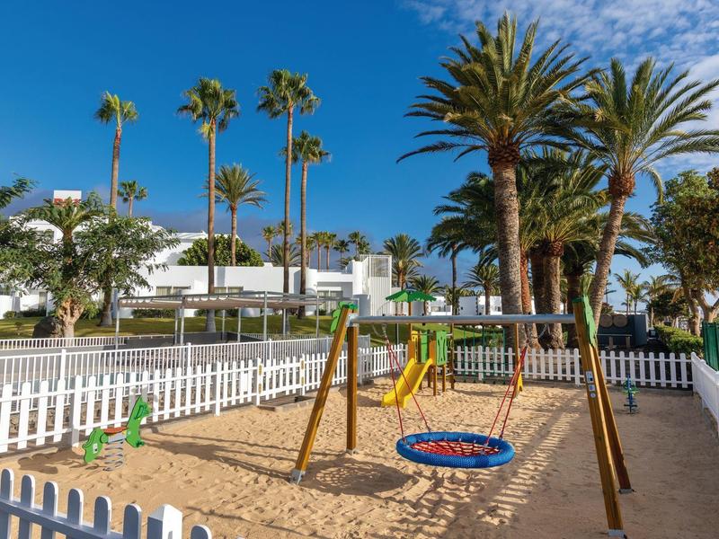 Area giochi per bambini con altalene circondata da palme e edifici bianchi.