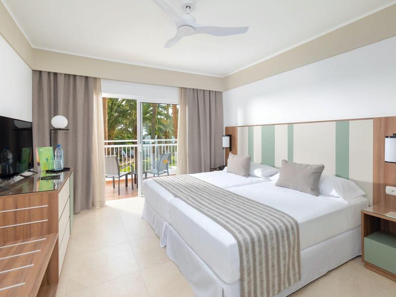 Camera d'albergo luminosa con letto matrimoniale, TV a schermo piatto e balcone con vista sulle palme.