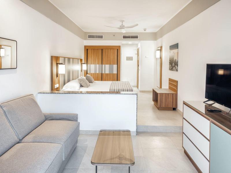 Camera d'hotel moderna con letto matrimoniale, divano, TV a schermo piatto e mobili in legno chiaro.