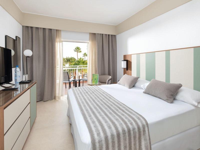 Camera d'hotel luminosa con letto matrimoniale, TV, balcone e vista sulle palme.