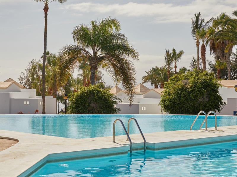 Piscina esterna dell'hotel circondata da palme e vegetazione sotto un cielo nuvoloso.