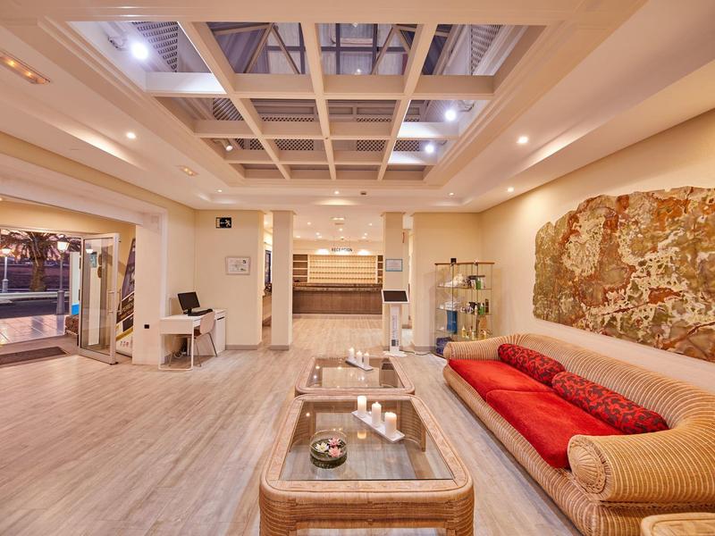 Ampio atrio d'hotel con area salotto, pareti decorate e pavimento in legno sotto una luce intensa.