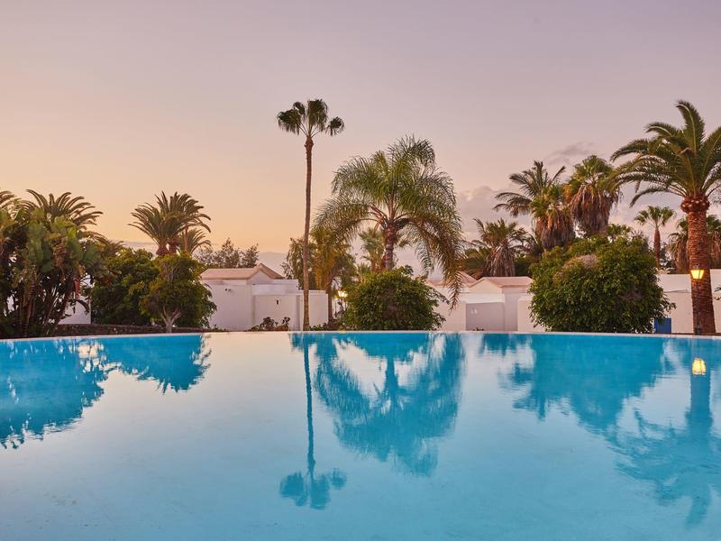 Piscina calma che riflette palme al tramonto in un ambiente di resort tropicale.