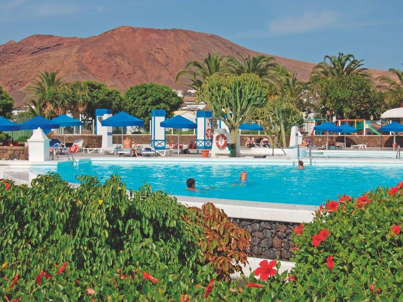 Piscina del resort con ombrelloni blu, sdraio, palme e montagna sullo sfondo.