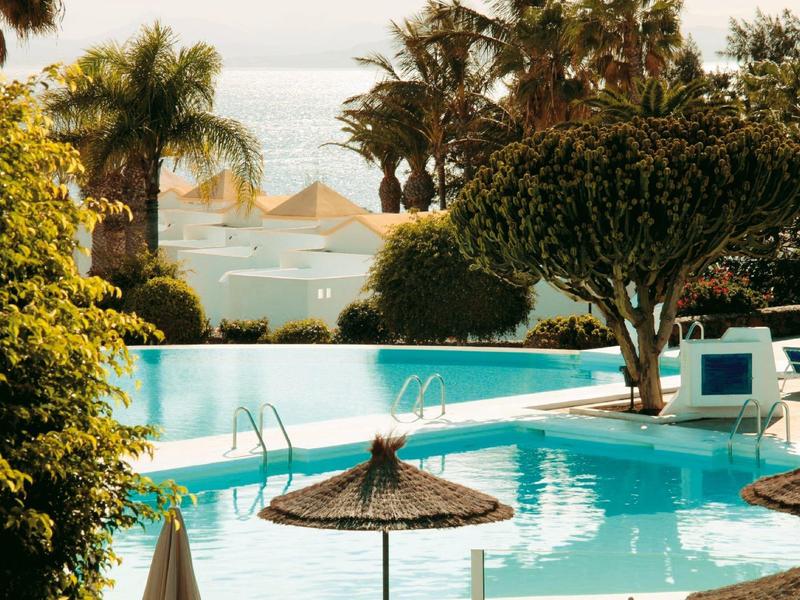 Area piscina dell'hotel con acqua blu, ombrelloni e palme vicino al mare.
