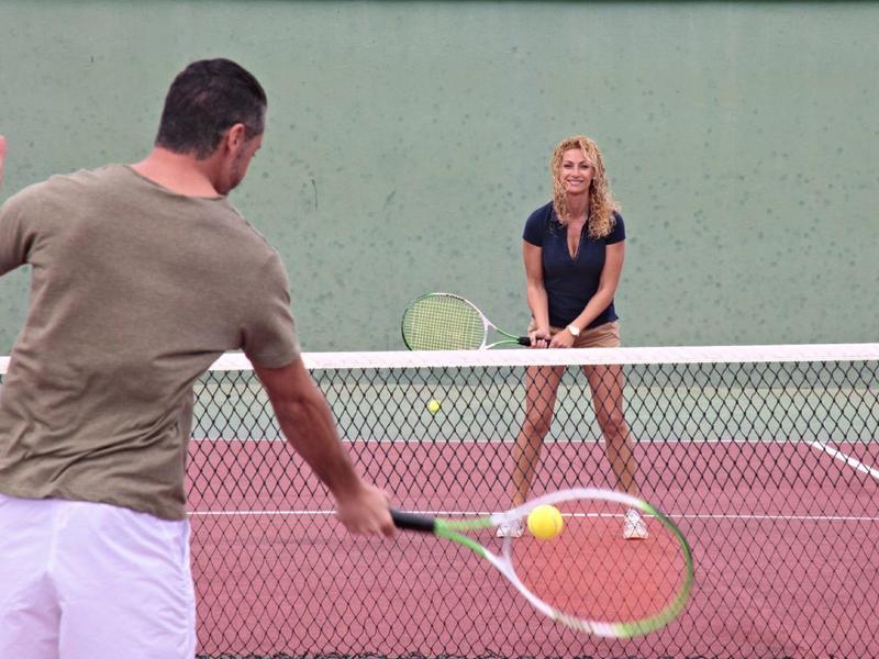 Due persone giocano a tennis su un campo rosso con uno sfondo verde.