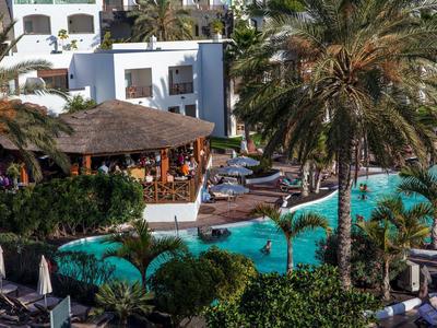 Piscine d'hôtel avec des palmiers, des parasols et des clients sous un toit de chaume en vacances.