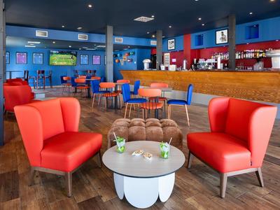 Espace bar d'hôtel moderne avec fauteuils rouges, chaises colorées et sol en bois.