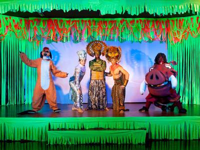 Stage performance with five characters from "The Lion King" in front of a green jungle backdrop.