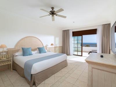 Chambre d'hôtel lumineuse avec lit double, ventilateur, balcon et vue sur la mer.