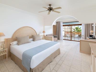 Chambre d'hôtel lumineuse avec grand lit, ventilateur de plafond et balcon avec vue sur l'eau.
