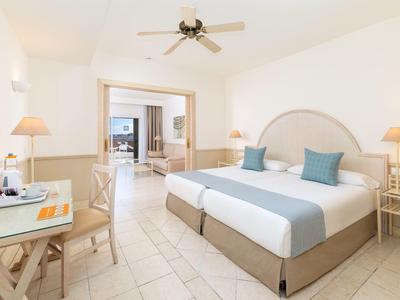 Chambre d'hôtel lumineuse avec deux lits simples, ventilateur de plafond, bureau et vue sur le salon.