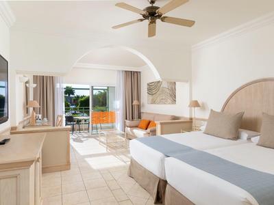 Chambre d'hôtel lumineuse avec lit double, ventilateur de plafond et balcon donnant sur la piscine.