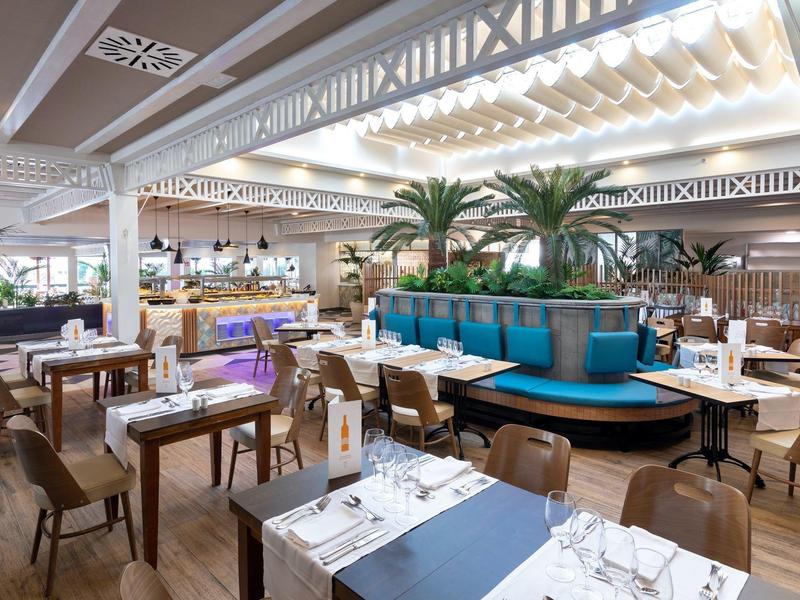 Restaurant moderne avec des tables en bois, des chaises et des palmiers en décoration intérieure.