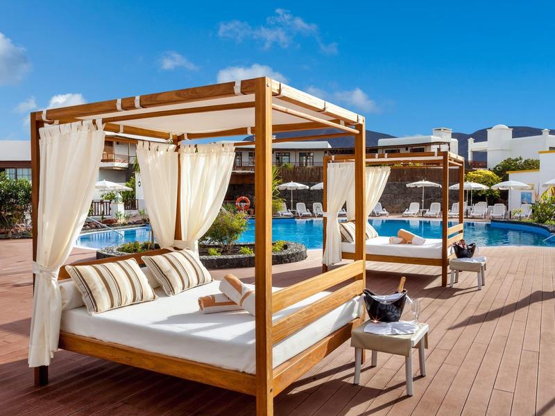 Lits en bois luxueux avec rideaux blancs à côté d'une piscine par une journée ensoleillée.