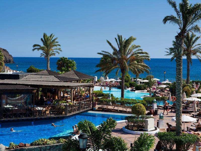 Piscine d'hôtel avec palmiers et vue sur la mer, chaises longues et parasols sous un ciel bleu