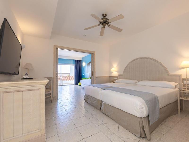 Chambre d'hôtel lumineuse avec lit double, TV écran plat, ventilateur de plafond et vue sur le balcon.
