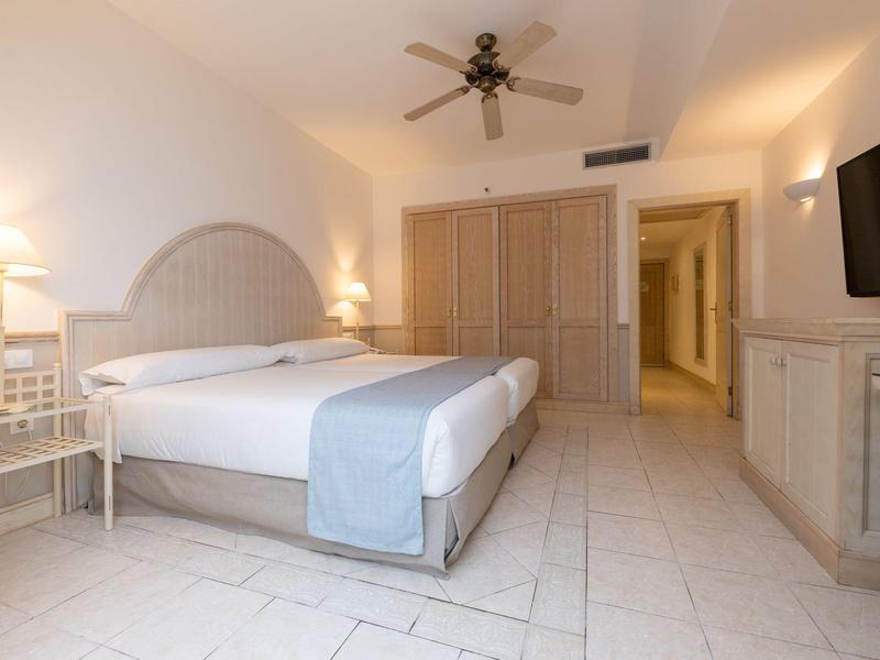 Chambre d'hôtel lumineuse avec lit double, ventilateur de plafond et télévision à écran plat au mur.