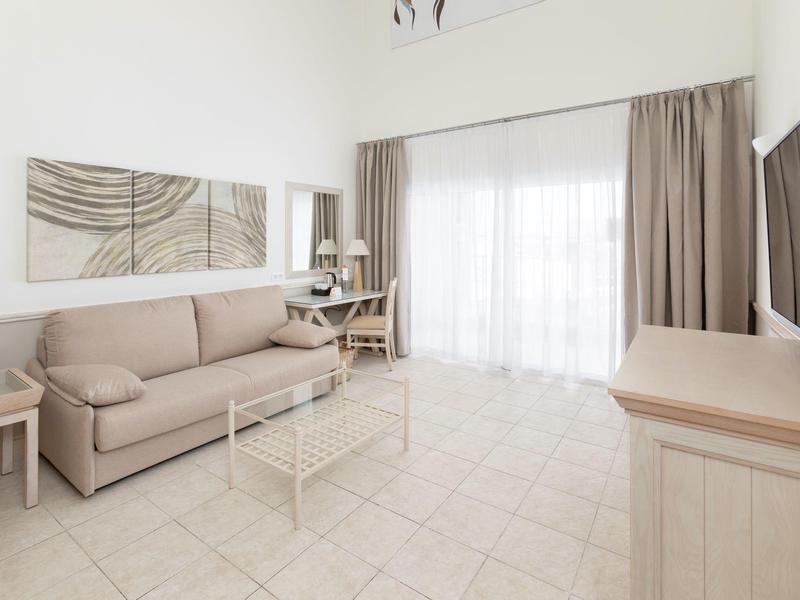 Chambre d'hôtel lumineuse avec canapé, table en verre, bureau et grande fenêtre avec rideaux.