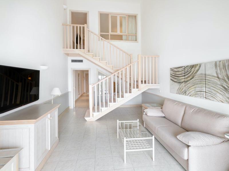 Salon lumineux avec mobilier blanc, canapé, TV et escalier menant à l'étage