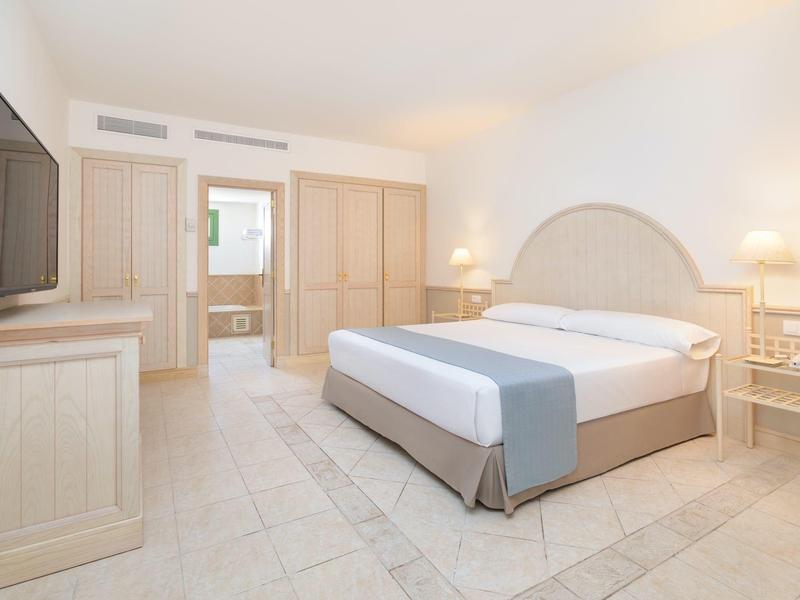 Chambre d'hôtel lumineuse avec grand lit double, armoire et télévision sur un sol carrelé clair.