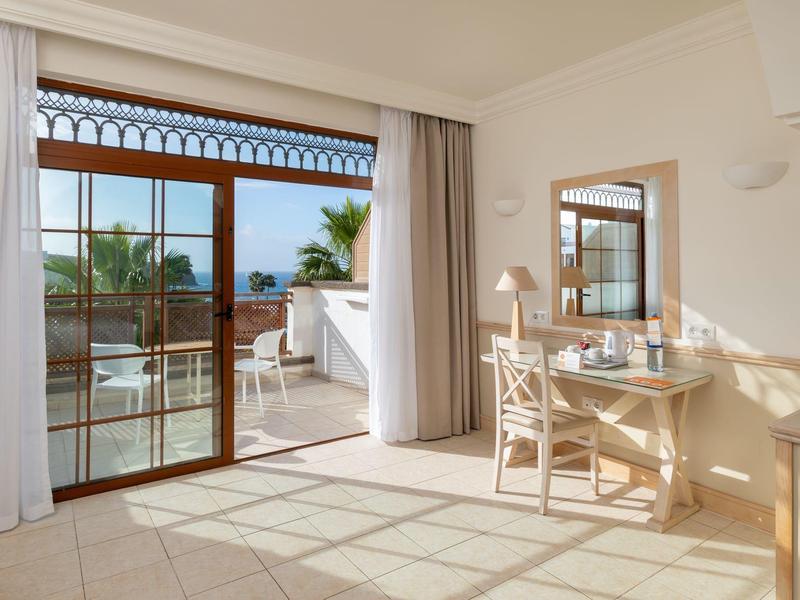 Chambre d'hôtel lumineuse avec bureau et grande fenêtre donnant sur une terrasse avec vue sur des palmiers par temps ensoleillé