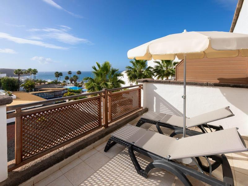 Balcon avec chaise longue, parasol et vue sur des palmiers et la mer en arrière-plan.