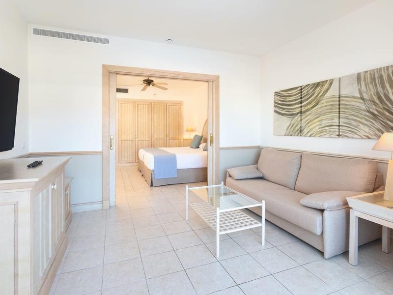 Salon lumineux avec canapé, table basse, télévision et accès ouvert à une chambre.