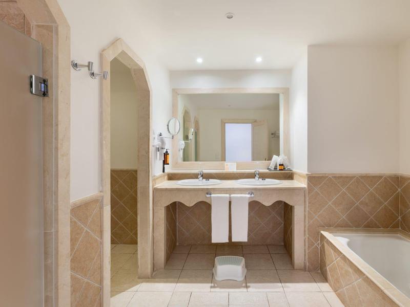 Salle de bain lumineuse avec grand miroir, lavabo, baignoire et murs carrelés beiges.