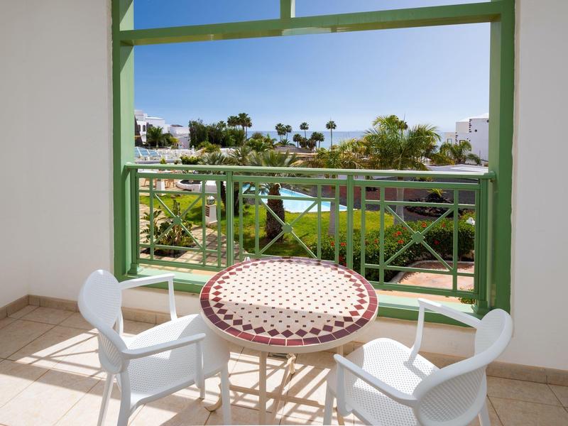 Balcon avec table et deux chaises donnant sur une piscine et des palmiers dans une station balnéaire.