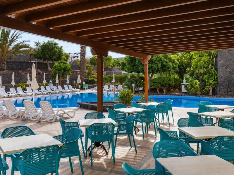 Terrasse couverte avec tables et chaises à côté d'une piscine et de transats dans un hôtel.