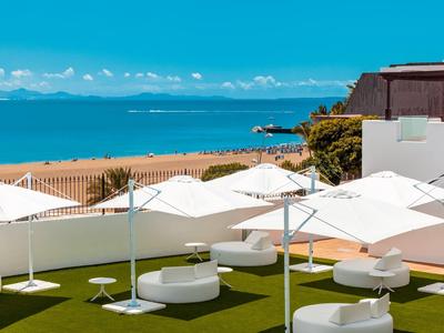 Modernes Strandhotel mit weißen Sonnenschirmen, runden Sitzmöbeln und Blauem Meer im Hintergrund.