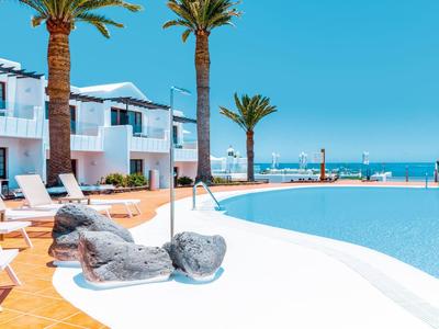 Modernes Hotel mit Pool, Sonnenliegen, Palmen und Blick aufs Meer bei klarem Himmel.