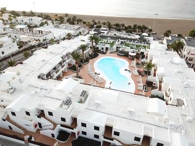 Blick auf weißes Hotel mit blauem Pool, Palmen und Strand im Hintergrund bei klarem Himmel.