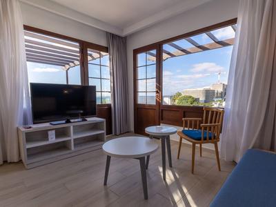 Hell eingerichtetes Zimmer mit Holzfenstern, Fernseher, runden Tischen und Stuhl mit blauem Sitz.