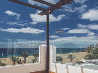 Terrasse mit orangefarbenen Fliesen, Glasgeländer und Aussicht auf Strand und blauen Himmel.