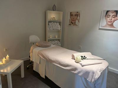 Hell eingerichteter Raum mit Massageliege, Handtüchern, Kerzen und sanfter Beleuchtung.