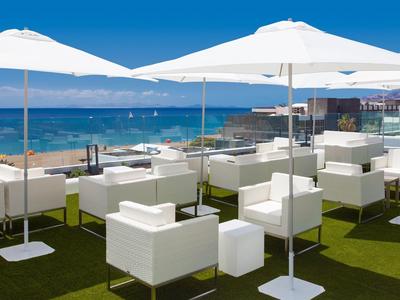 Strandterrasse mit weißen Sesseln, Tischen und Sonnenschirmen vor blauem Meer und klarem Himmel.