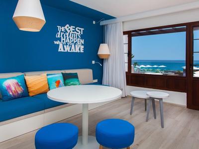 Helles Wohnzimmer mit blauer Wand, rundem Tisch, blauen Hockern und Meerblick durch großes Fenster.