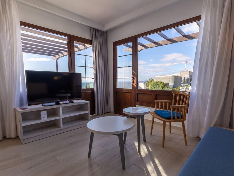 Camera d'hotel luminosa con TV, due tavoli e vista su un balcone alla luce del giorno.