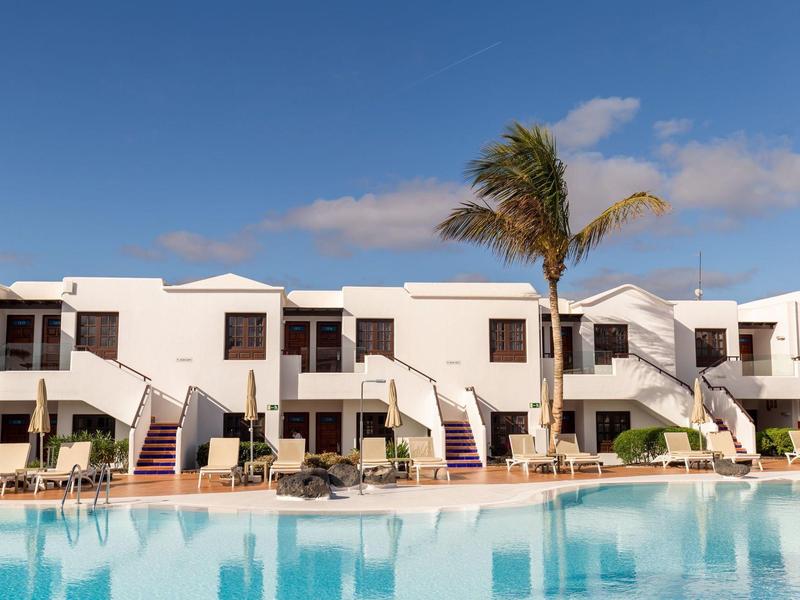 Edifici bianchi con terrazze e piscina in primo piano sotto un cielo limpido e blu.