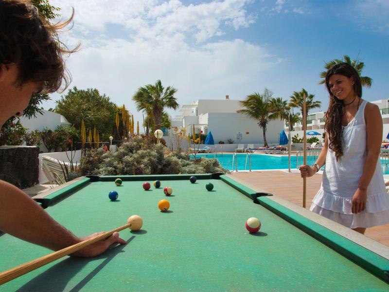 Due persone giocano a biliardo all'aperto accanto a una piscina in un resort vacanze con palme.