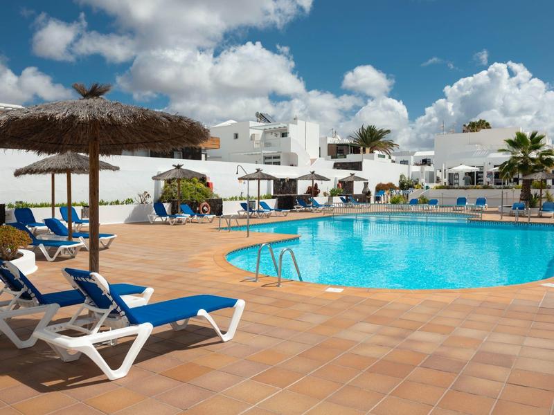Area piscina con lettini e ombrelloni davanti a edifici bianchi sotto un cielo azzurro.