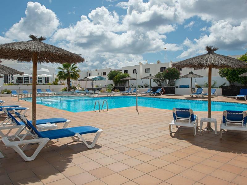 Area piscina con lettini blu e ombrelloni di paglia di fronte a case vacanze bianche.